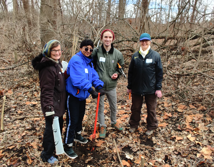 Four people smiling at the camera as they prepare to plant a tree in the woods.